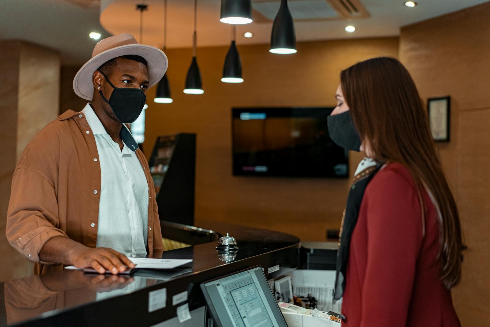 A man and woman in face masks checking in at a hotel reception, emphasizing safety protocols.